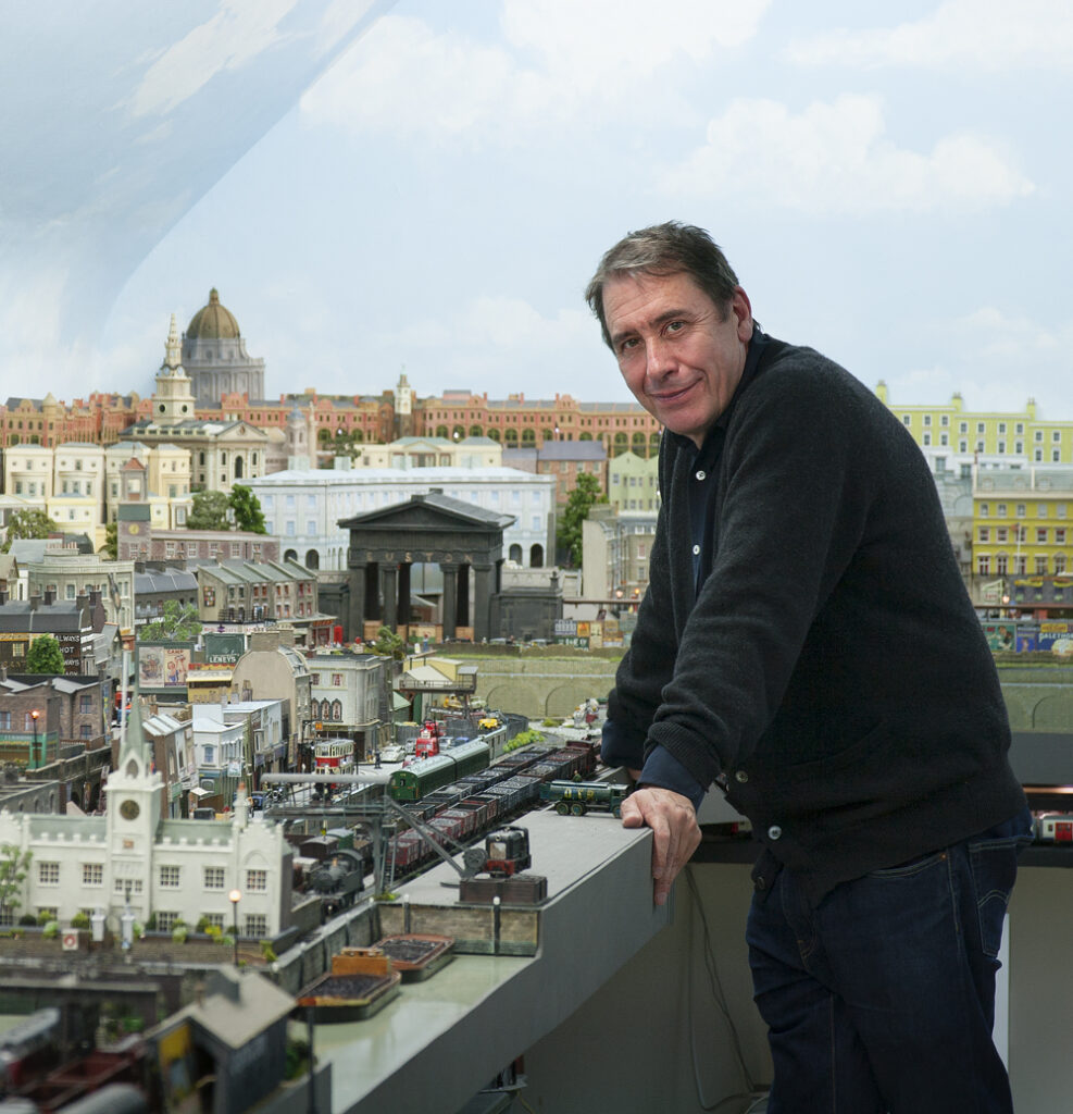 Man leans over train layout, looking at camera.