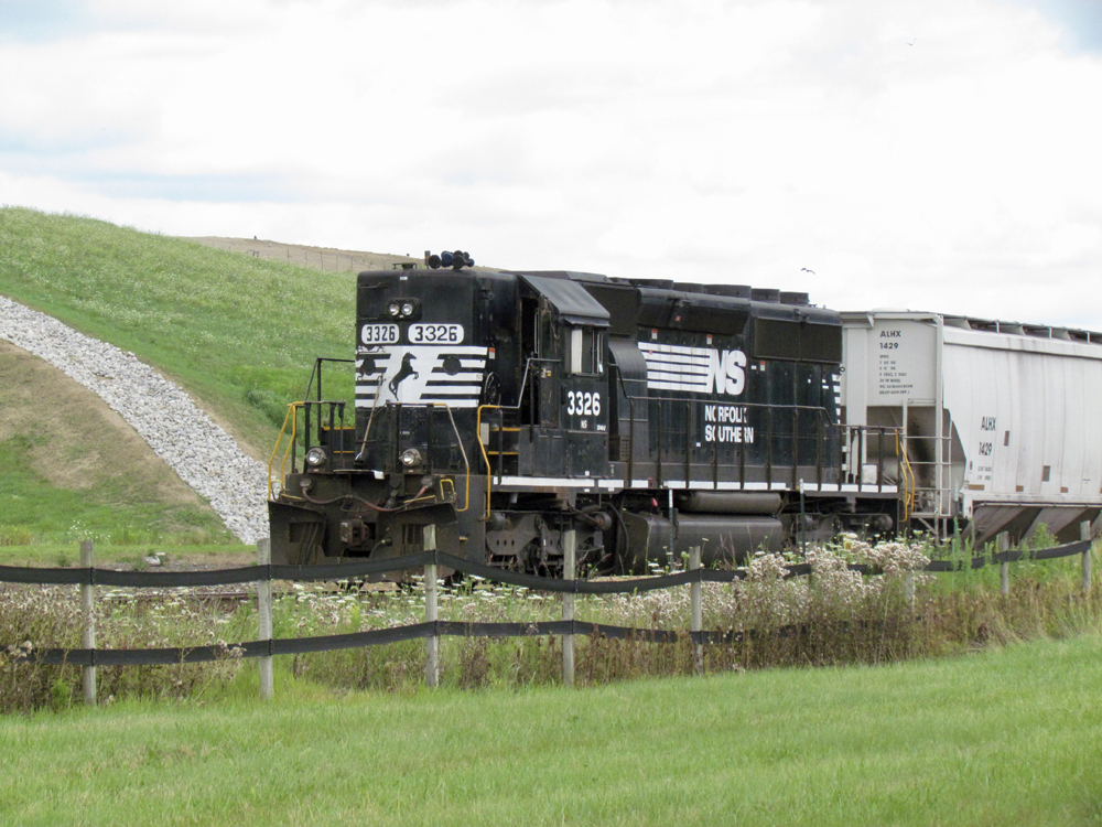 Black train engine leads train past fences and a reservoir