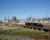 Black steam locomotive pulling 12-car passenger excursion train past an oil refinery.