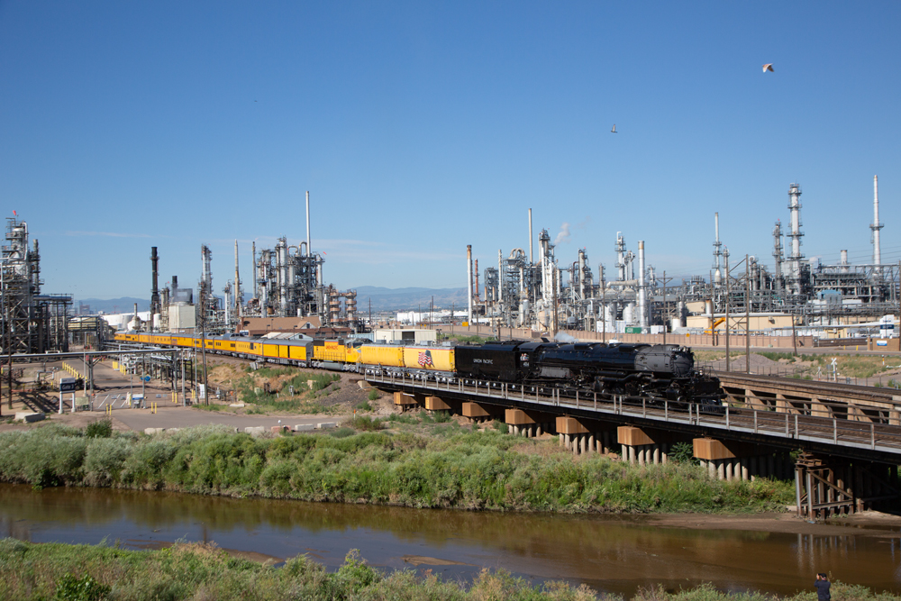 Black steam locomotive pulling 12-car passenger excursion train past an oil refinery.