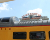 Dome and roof of a yellow and gray dome car with train station sign in background.