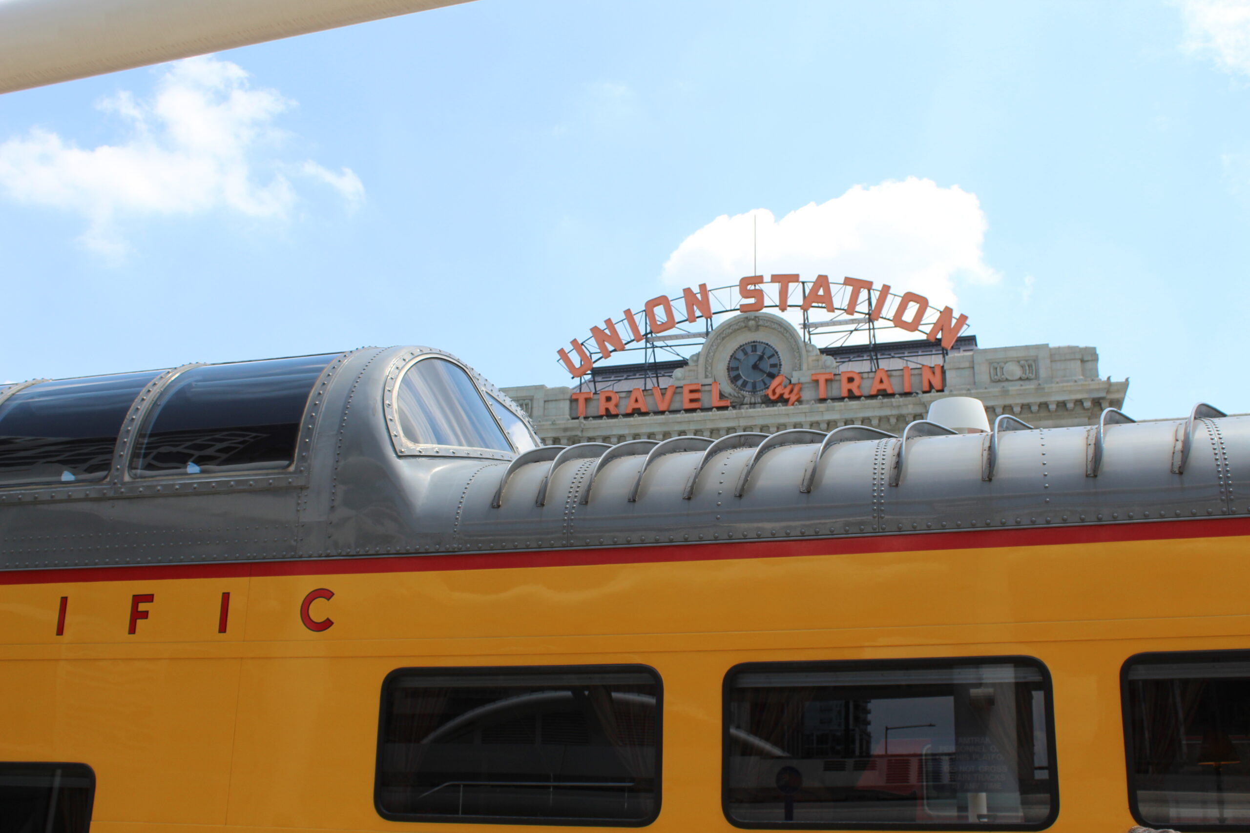 Dome and roof of a yellow and gray dome car with train station sign in background.