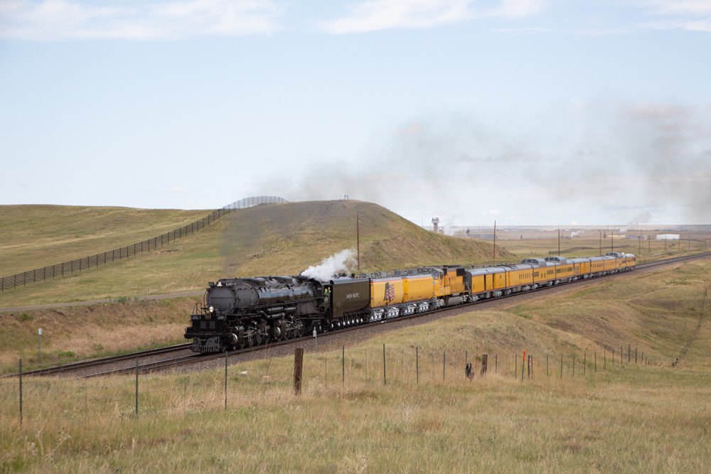 Black steam locomotive pulling 12-car passenger excursion train through rolling hills.