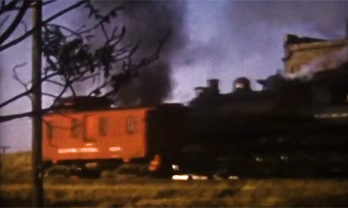 Old home-movie image of a black steam locomotive pushing a red caboose.