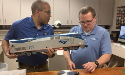 Two men at a table, one holding a large-scale model train locomotive.