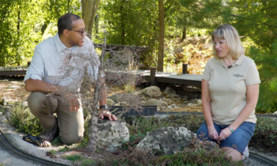 Man and woman kneel, talking in a garden railroad.