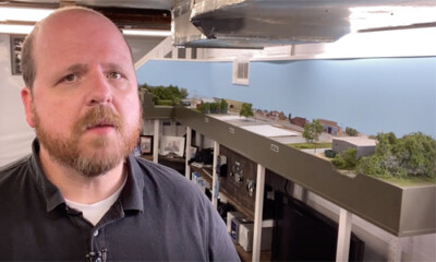 Man looking at camera with model train layout behind him.