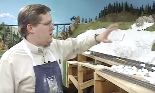 Man gesturing with an arm over an unfinished model railroad scene.