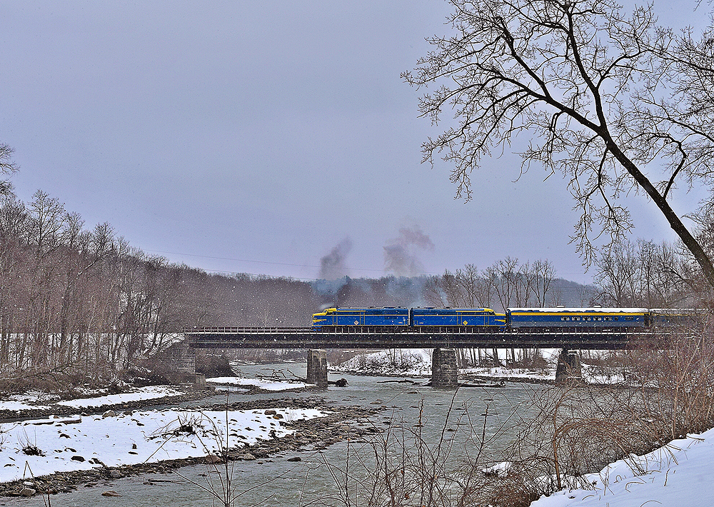 Blue and yellow passenger train cross a bridge in a snowy scene.