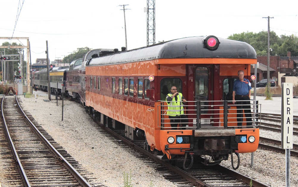 Train with orange and maroon observation car at end backs toward camera