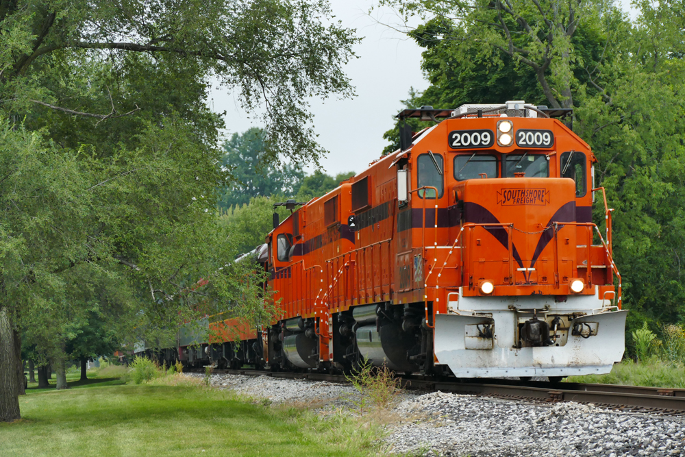 Two orange locomotives with passenger train in tree-lined area