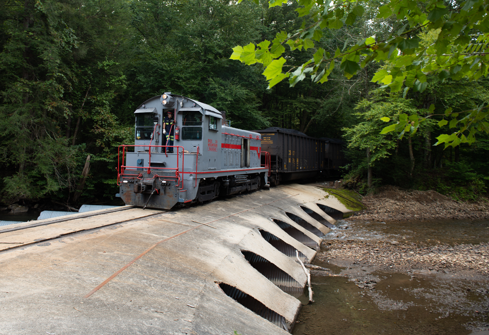 Gray end-cab switcher crossing culvert with coal hoppers
