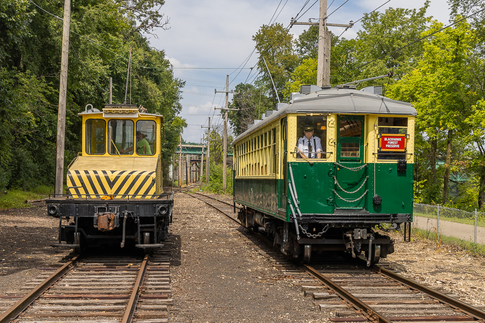 Yellow steeplecab electric locomotive and green and yellow transit car on adajcent tracks