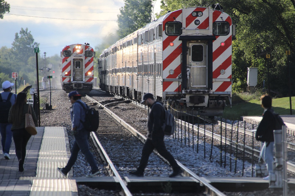 People walk across railroad tracks as trains meet in background