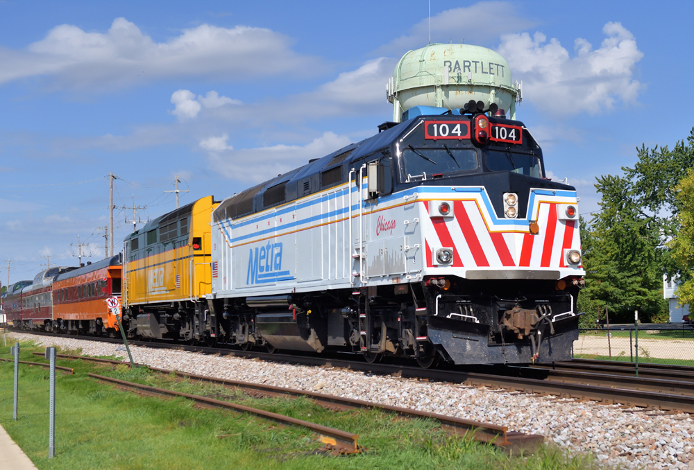 White and gray locomotive leads yellow and green locomotive and string of mismatched passenger cars