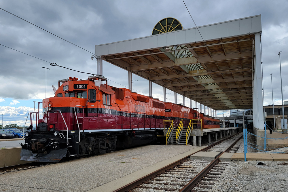 Two orange and maroon locomotives and one passenger cars under shed at two-track station