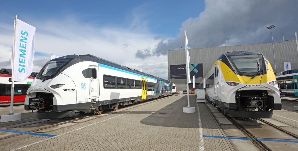 Two passenger trains, one white and blue, one white and yellow, on display tracks outside exhibition hall.