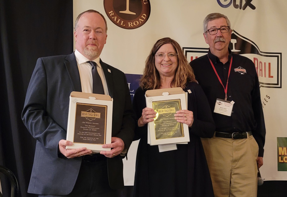 Three people standing on platform, two holding award plaques