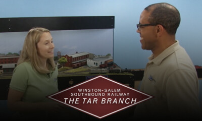 Man and woman speaking in front of a model train layout with logo.