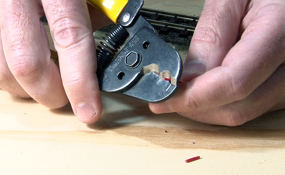 Hands holding a gray metal wire cutter with its blades wrapped around a small red wire.
