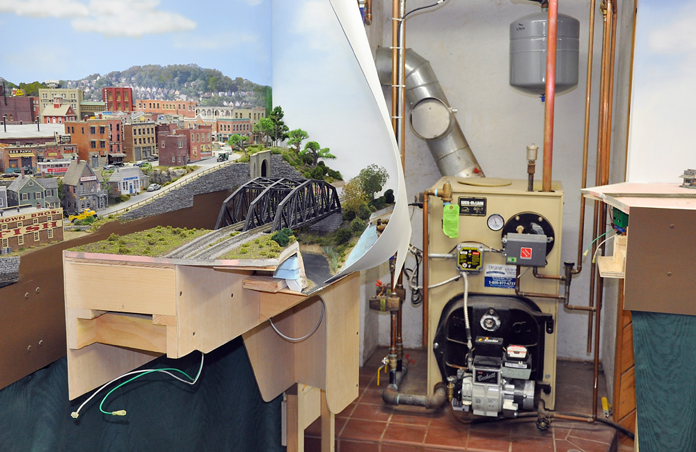 A section of a train layout built on a swinging gate stands open, giving access to a home furnace behind it