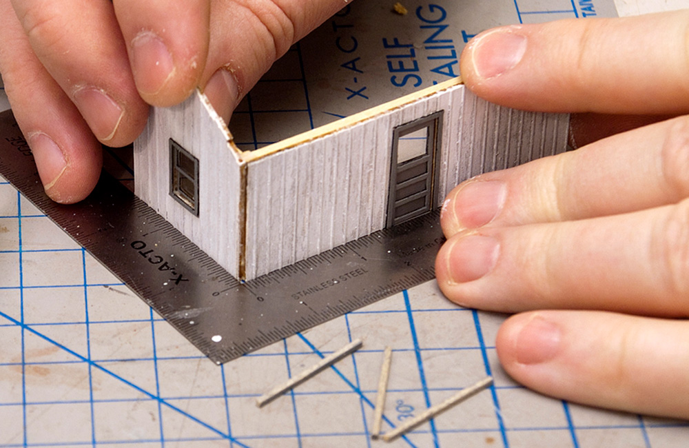 Two walls of a small wooden structure kit are glued together, aligned by a small machinist’s square