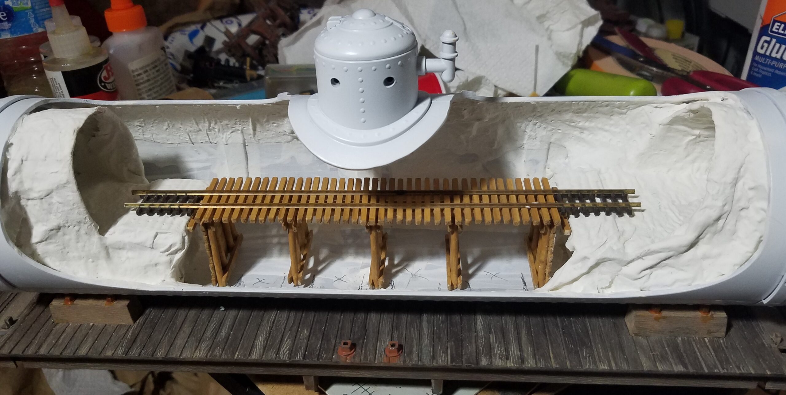 A large scale tank car model cut open with white plaster scenery and N scale track on a wooden trestle