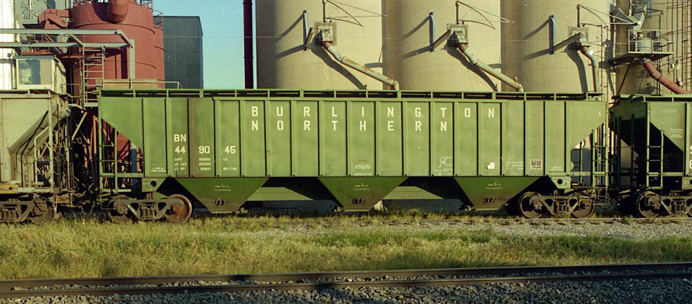 Photo of green covered hopper in front of grain elevator on sunny day