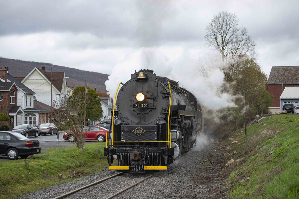 Steam locomotive passes through town under cloudy skies