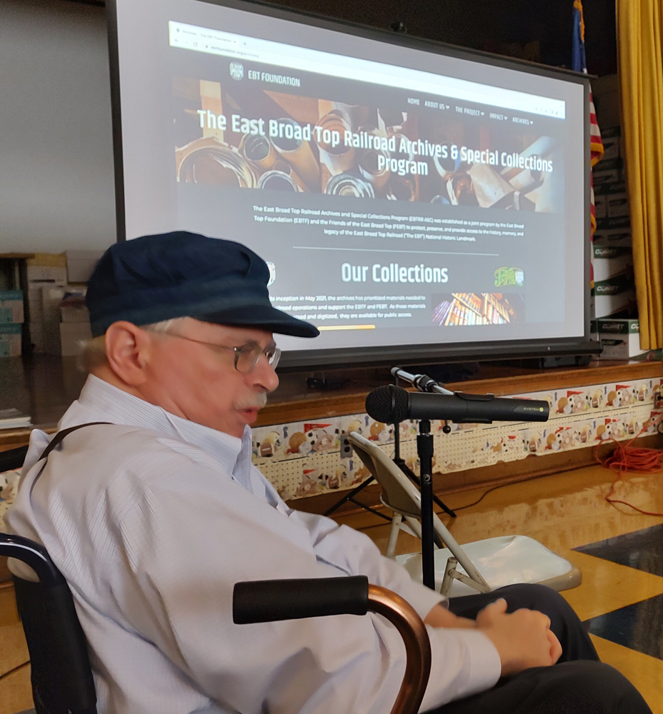 Man in chair speaking into microphone with image on screen in background