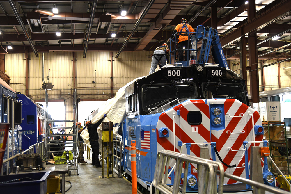 Front view of blue locomotive with red and white nose stripes in shop building