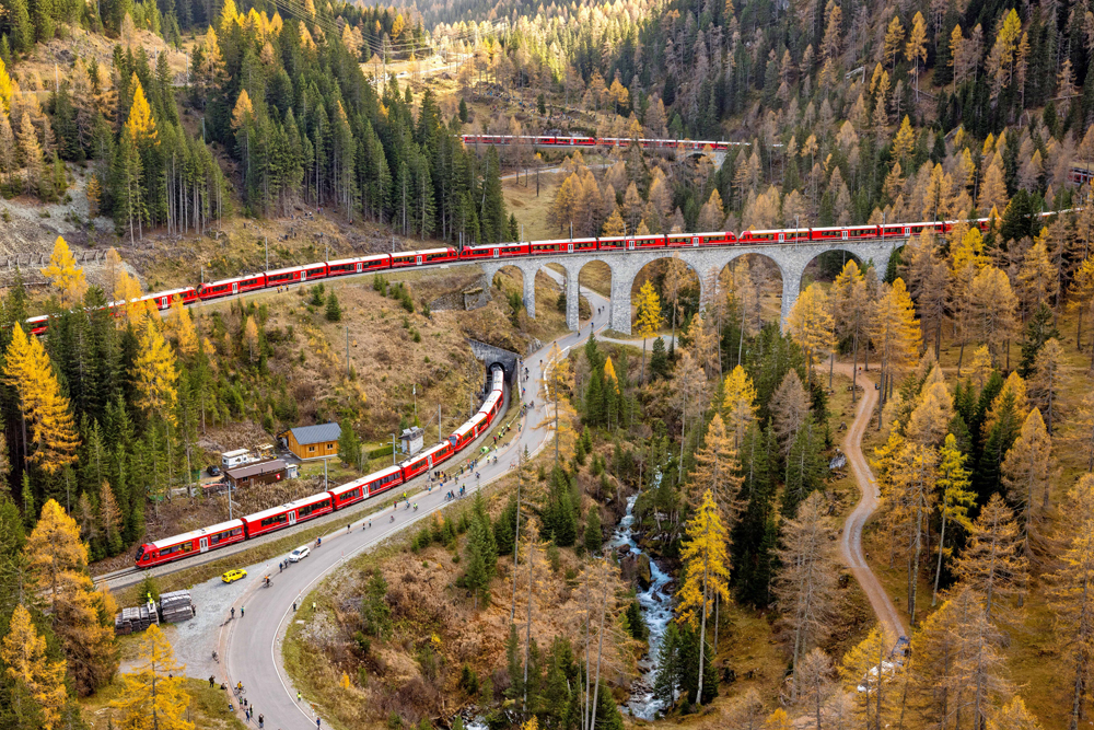 Long red train on several levels of tracks, with the front coming out of the tunnel