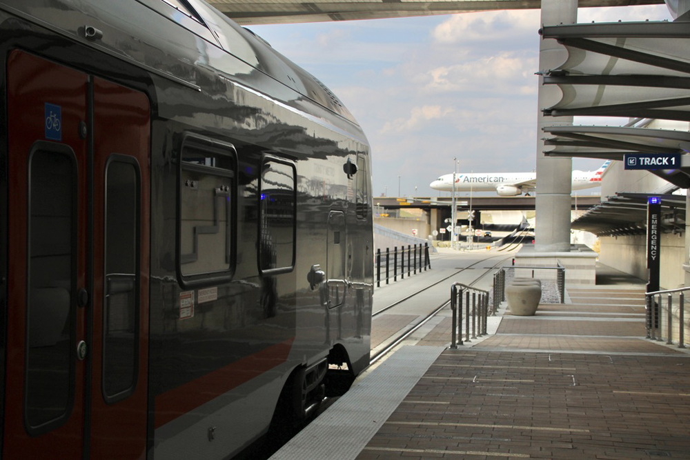 Diesel multiple-unit train in station with airplane crossing bridge in background. Traveling Dallas-Fort Worth by rail.