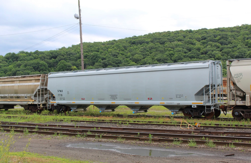 A new light gray, four-bay, cylindrical covered hopper is seen in a rail yard