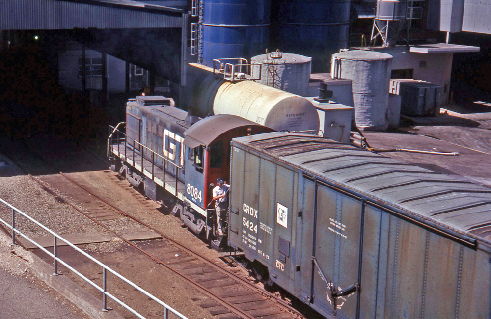 A dark blue switch engine pulls a boxcar past a cluster of tanks and silos