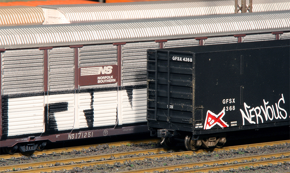 an enclosed auto rack sits next to a black boxcar on an adjacent track, both with graffiti on them