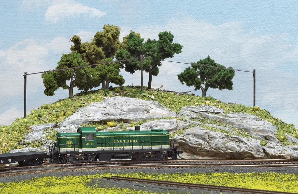 A green-and-white Alco diesel passes by a rock cut in a hill topped with trees and line poles