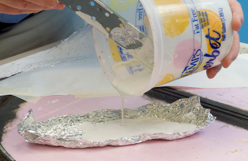 Plaster is poured from a plastic bucket into a mold of crumpled aluminum foil