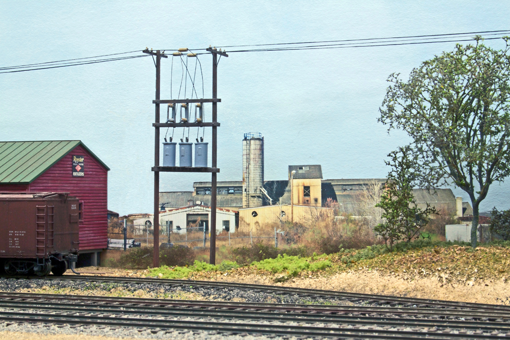 A foundry is shown in the background against a blue sky backdrop, with a transformer bank and tree in he foreground along model railroad track