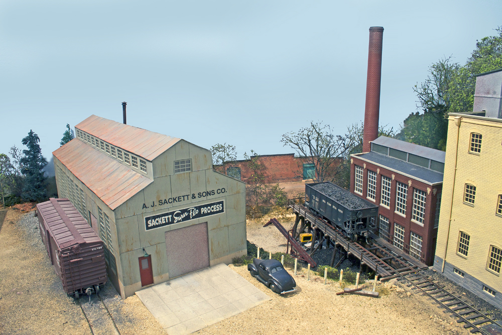 A warehouse and a factory are shown along a coal trestle and a stretch of model railroad track, with a coal car and a freight car along the structures