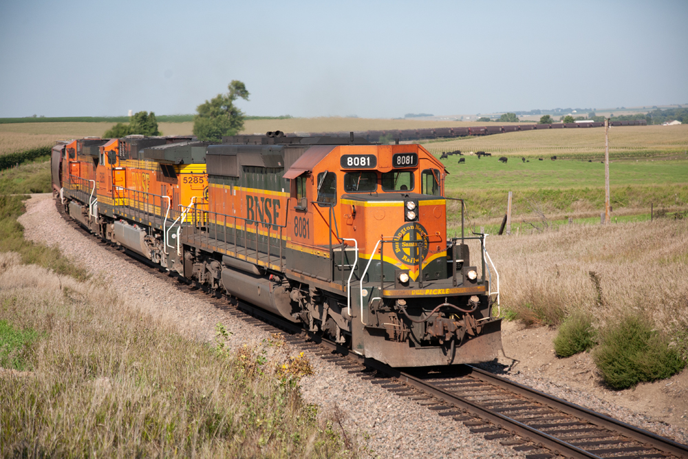 green and orange freight train passing open farm fields