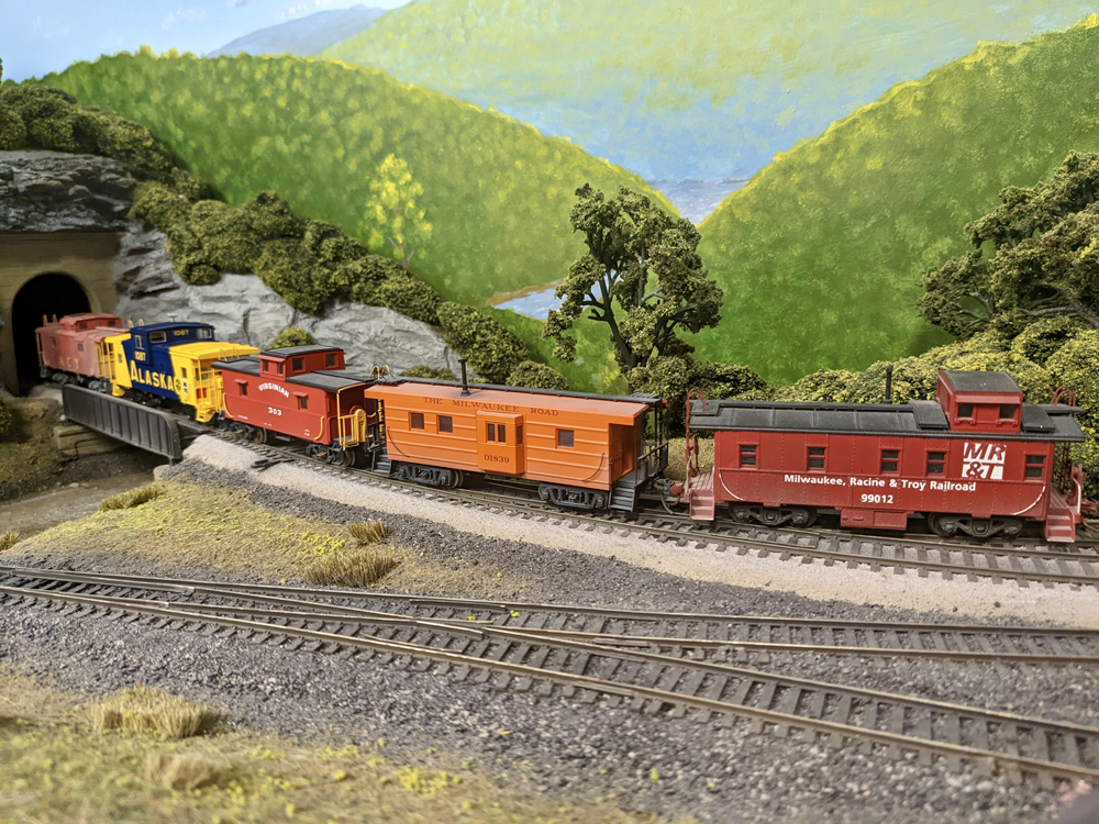 Five colorful model cabooses go into a tunnel on a mountainous train layout