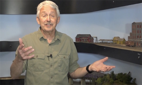 Man in front of a model train layout, gesturing.