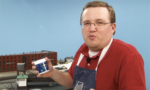 Man holding a container of modeling product in front of a model train layout.