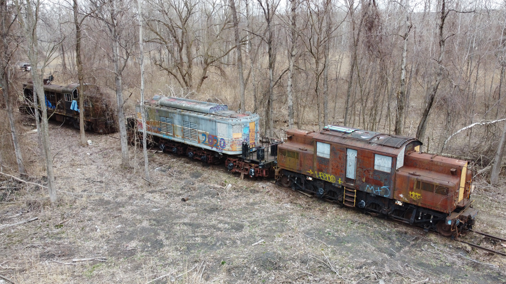 Drone view of two electric locomotives and one diesel sitting vandalized in woods