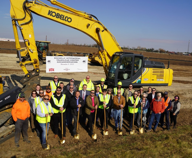 People with shovels pose in front of earthmoving equipment