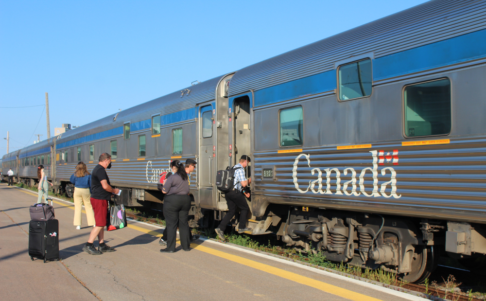 Passengers boarding stainless steel vintage passenger cars with blue stripes above windows and "Canada" lettering.