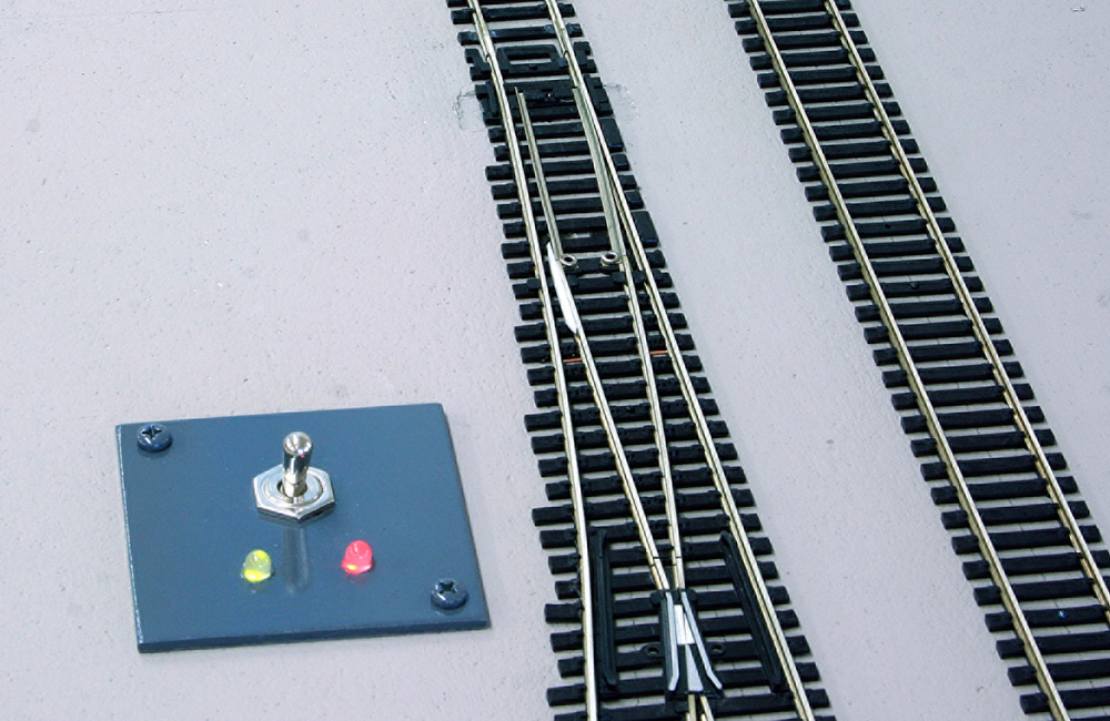 A small panel next to a model railroad turnout has a toggle switch and two red and green LEDs