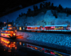 A train decorated with Christmas lights rolls along a riverbank at night, past a brightly lighted riverboat