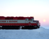 A red-and-white diesel in profile pushes snow off the tracks as the sun rises behind it